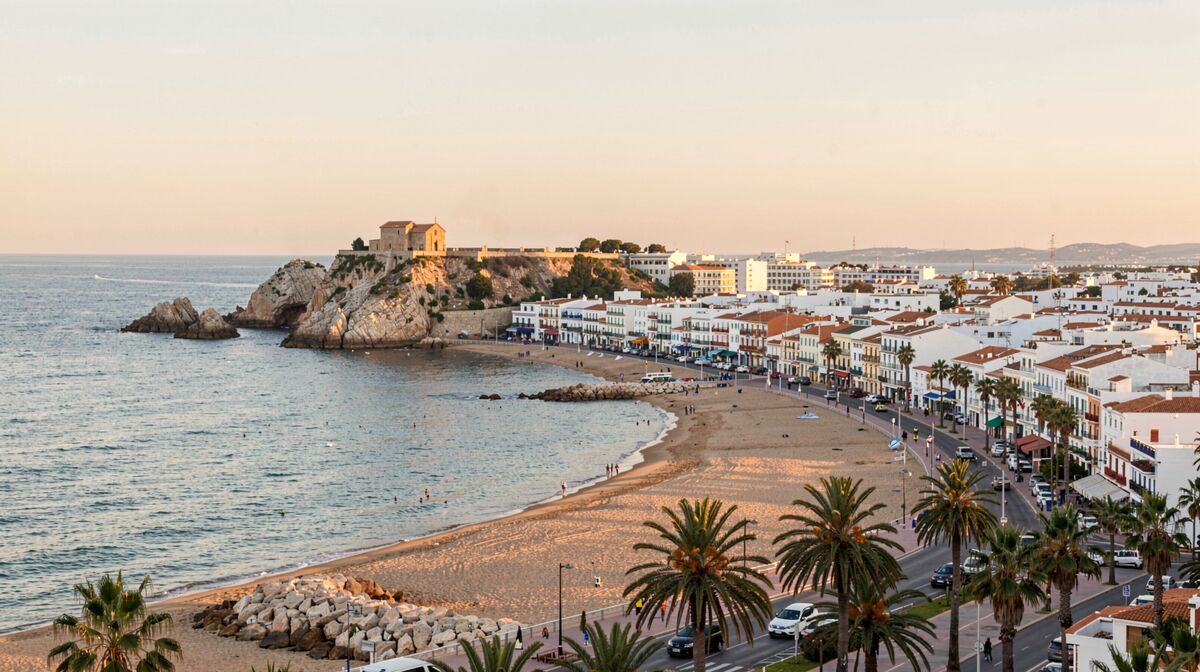 Vista de la villa de Sitges al atardecer, con la iglesia sobre la roca, la playa curva y el paseo de palmeras