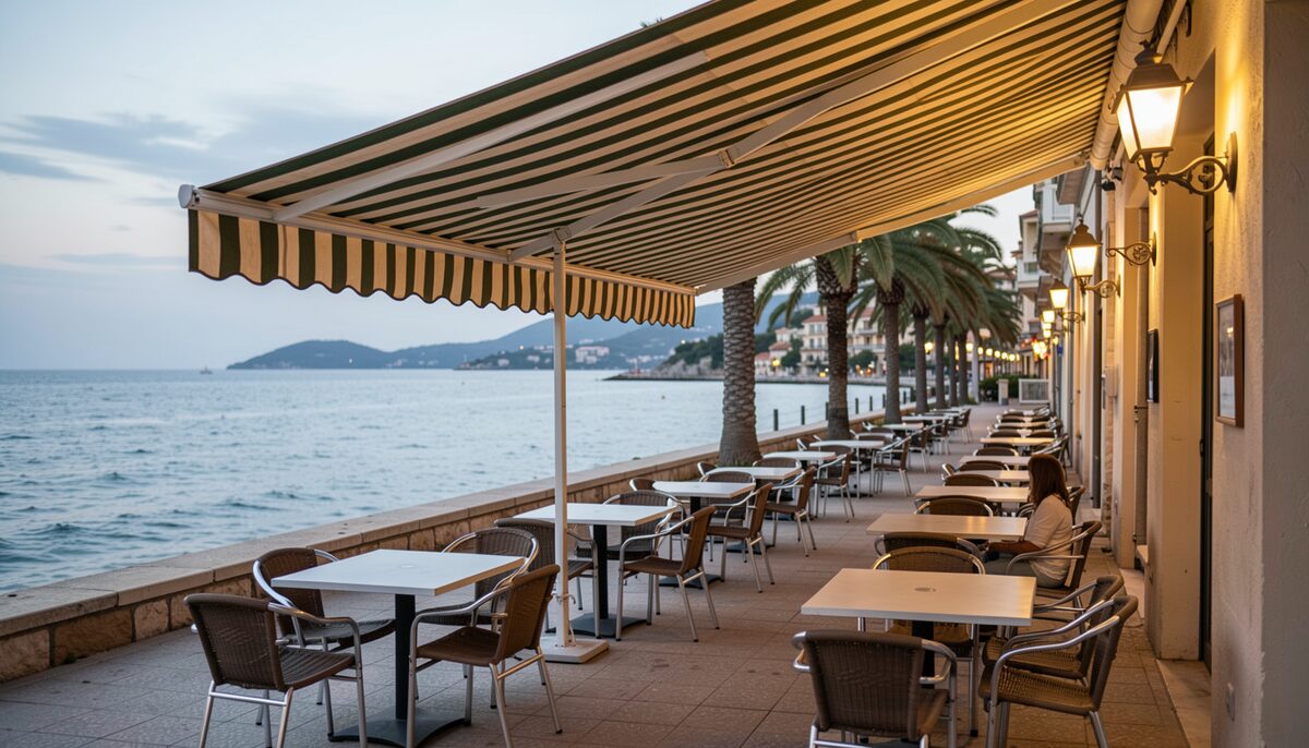 Terraza con mesas vacías bajo un toldo de rayas frente al mar, en un paseo con palmeras